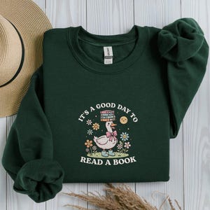 Può includere: Felpa verde scuro con una grafica di un'oca con fiocco rosa e stivali, che trasporta libri. Il testo "IT'S A GOOD DAY TO READ A BOOK" è stampato. Il design include fiori e un sole sorridente. Un cappello di paglia è visibile.