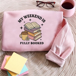 May include: A pale pink sweatshirt featuring the phrase "MY WEEKEND IS FULLY BOOKED" in black, with a graphic of books and a coffee cup. The design includes floral elements. The sweatshirt is folded on a woven surface.