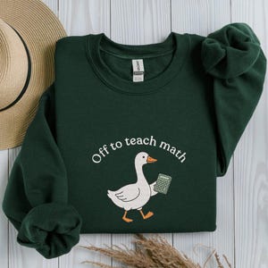 May include: A dark green sweatshirt featuring the phrase "Off to teach math" in a circular design. A cartoon goose holding a calculator is in the center. The sweatshirt is made of a soft material.
