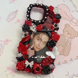 May include: A decorative phone case featuring a photo of a person. The case is adorned with red and black roses, pearls, and a white butterfly. The text "art shaker" is visible on the case.