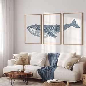 May include: Three framed prints of a blue whale illustration. The whale is depicted in a line art style with a white background. The prints are hung on a white wall above a white sofa with blue throw blankets.