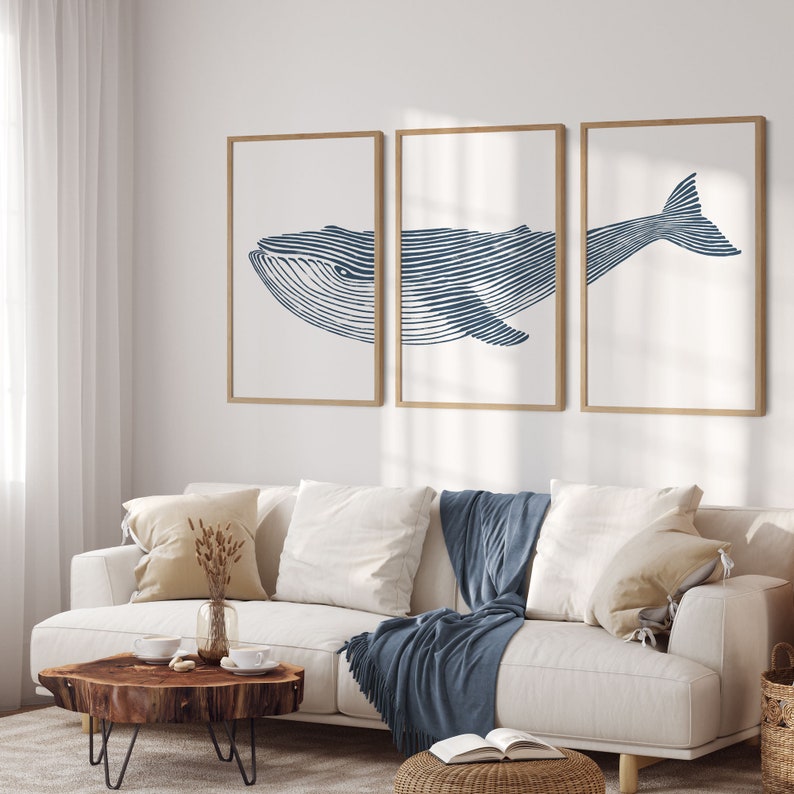 May include: Three framed prints of a blue whale illustration. The whale is depicted in a line art style with a white background. The prints are hung on a white wall above a white sofa with blue throw blankets.