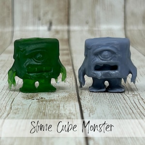 May include: Two cube-shaped monster figurines. One is translucent green, the other is gray. Both have a single eye, a wide mouth, and small arms and legs. The text "Slime Cube Monster" is written in cursive at the bottom.