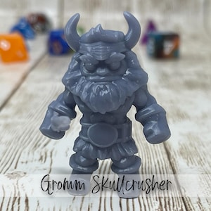 May include: A gray Gromm Skullcrusher figurine with a horned helmet, beard, and belt. The miniature figure is standing, with other colorful dice in the background. The text "Gromm Skullcrusher" is written at the bottom of the image.