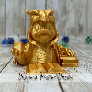 May include: A golden-colored Dungeon Master Ducky figurine. The duck is wearing a hooded robe and holding a geometric shape in one hand and a stack of books in the other. The text "Dungeon Master Ducky" is at the bottom.