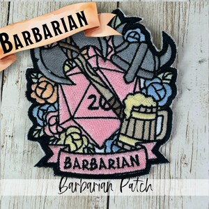May include: An embroidered patch featuring a pink twenty-sided die, a grey horned helmet, a battle axe, a wooden beer mug, and colorful flowers. A banner at the bottom displays the text BARBARIAN in black, with a matching banner at the top.