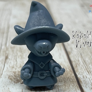 May include: A blue 3D printed figurine of a kobold wizard wearing a pointed hat and a robe. The figurine is standing on a wooden surface. The text "Kobold Wizard" is written in black on the right side of the image.