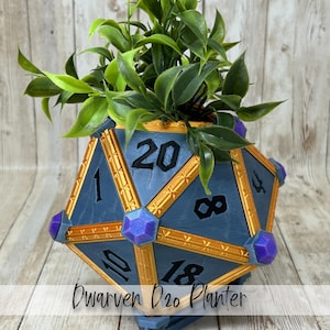 May include: A blue and gold D20 planter with a geometric design, featuring numbers and purple accents. The planter is filled with green leafy plants. The text "Dwarven D20 Planter" is visible at the bottom.