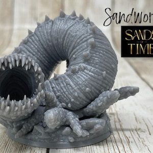 May include: A gray 3D printed sandworm miniature with a large open mouth and a textured body. The miniature is on a base with a small rocky texture. The text "Sandworm" and "Sands of Time" is visible in the image.