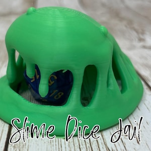 May include: A green slime dice jail with a blue and gold dice inside. The slime jail is shaped like a blob with holes for the dice to fall through. The text "Slime Dice Jail" is written below the jail.