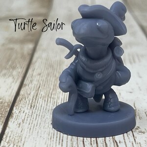May include: A gray figurine of a turtle dressed as a sailor, complete with a hat, a sword, and a walking stick. The figurine is standing on a round base. The text "Turtle Sailor" is written in cursive.