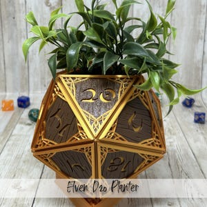 May include: A brown and gold Elven D20 planter with a faux plant. The planter is shaped like a twenty-sided die, with the numbers 1-20 visible. The planter has a gold trim and a wood-grain texture. The text "Elven D20 Planter" is at the bottom.