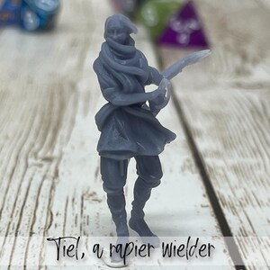 May include: A gray miniature figure of a person wielding a rapier. The figure is dressed in period clothing, with a scarf covering the lower face. The text "Tiel, a rapier wielder" is at the bottom of the image. Dice are in the background.