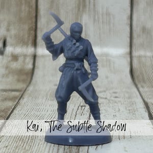 May include: A gray miniature figure of a ninja, holding two pickaxes. The figure is standing on a gray base. The text "Kai, The Subtle Shadow" is written across the bottom of the image.