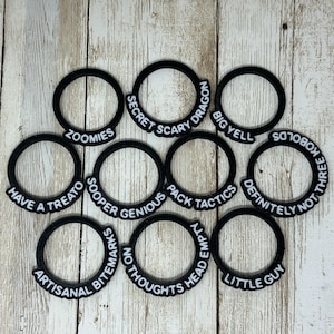 May include: A collection of black circular objects with various white text labels. The labels include phrases like "ZOOMIES", "SECRET SCARY DRAGON", and "BIG YELL". The objects are arranged on a light-colored wooden surface.