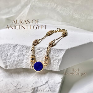 May include: Gold-toned bracelet featuring a deep blue circular stone at its center. The bracelet has intricate detailing with small, decorative elements and the text "AURAS OF ANCIENT EGYPT". The design evokes an ancient Egyptian aesthetic.