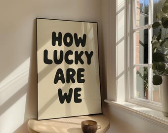 May include: A framed art print with the words "HOW LUCKY ARE WE" in bold, black letters on a cream background. The artwork is displayed on a small wooden table, with a coffee cup. A window and a potted plant are in the background.