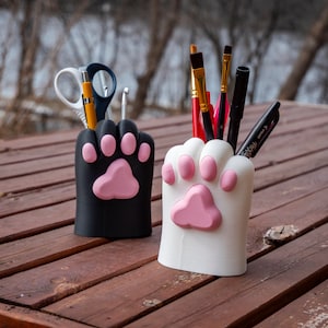 Op de afbeelding: Twee potloodhouders in de vorm van een kattenpoot, een zwart en een wit, met roze pootkussentjes. De zwarte houder bevat een schaar, een potlood en een haaknaald. De witte houder bevat verschillende kwasten en een zwarte stift.