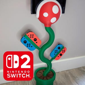 May include: A green, red, and white decorative Nintendo Switch game console holder shaped like a Piranha Plant. The holder has two blue and red Joy-Con controllers attached to the stem. The Nintendo Switch logo is visible.