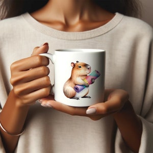 May include: White ceramic mug with a cartoon illustration of a capybara holding a rolled-up yoga mat. The capybara is brown with a pink nose and a white belly. The yoga mat is rainbow colored.