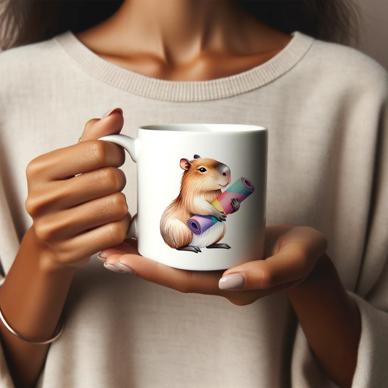 May include: White ceramic mug with a cartoon illustration of a capybara holding a rolled-up yoga mat. The capybara is brown with a pink nose and a white belly. The yoga mat is rainbow colored.