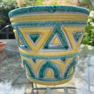 May include: A ceramic plant pot with a geometric design. The pot has a yellow and teal colour scheme with a repeating triangle pattern. The rim and base are teal, and the body is yellow with teal accents. Suitable for indoor or outdoor use, it measures approximately 15 cm in height.