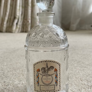 Op de afbeelding: Een doorzichtige glazen fles met een getextureerd oppervlak en een ronde stop. De fles heeft een etiket met de tekst "Eau de Cologne du Coq Guerlain 68, Champs-Elysees, 68 Paris".
