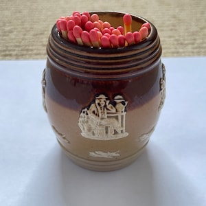 May include: A brown ceramic match holder with a white design of people and animals. The holder is filled with red and white matches.