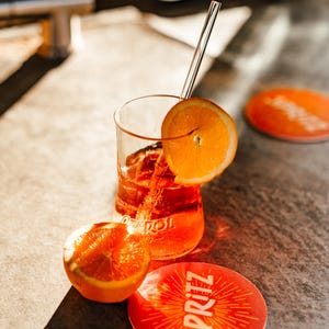 May include: A glass of orange-colored beverage with a slice of orange and a straw, sitting on a coaster with the word "SPRITZ" printed on it. There is another orange slice next to the glass.