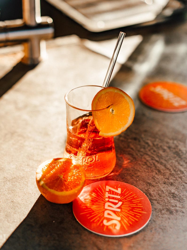 May include: A glass of orange-colored beverage with a slice of orange and a straw, sitting on a coaster with the word "SPRITZ" printed on it. There is another orange slice next to the glass.