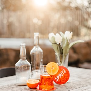 May include: A glass of orange juice with an orange slice garnish, sitting on a red coaster with the word "SPRITZ" printed on it. Two clear glass bottles and a vase of white tulips are in the background.