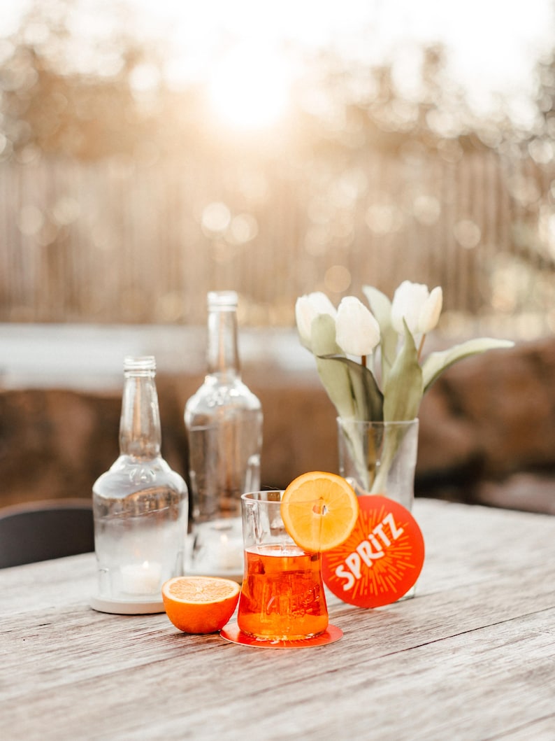 May include: A glass of orange juice with an orange slice garnish, sitting on a red coaster with the word "SPRITZ" printed on it. Two clear glass bottles and a vase of white tulips are in the background.