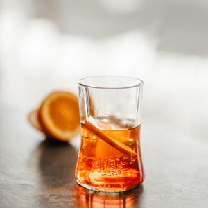 May include: A glass of Aperol Spritz with an orange slice, the glass has the Aperol logo and the year 1919 printed on it.
