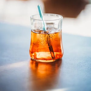 Könnte beinhalten: Ein Glas orangefarbener Flüssigkeit mit einem blau-weiß gestreiften Strohhalm. Das Glas hat ein Logo auf der Vorderseite und steht auf einer reflektierenden Oberfläche. Das Getränk scheint ein Cocktail oder ein Getränk zu sein.