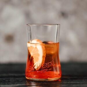 May include: A glass of Aperol Spritz with a slice of orange. The glass is clear and has the word "Aperol" printed on it. The drink is a bright orange color.