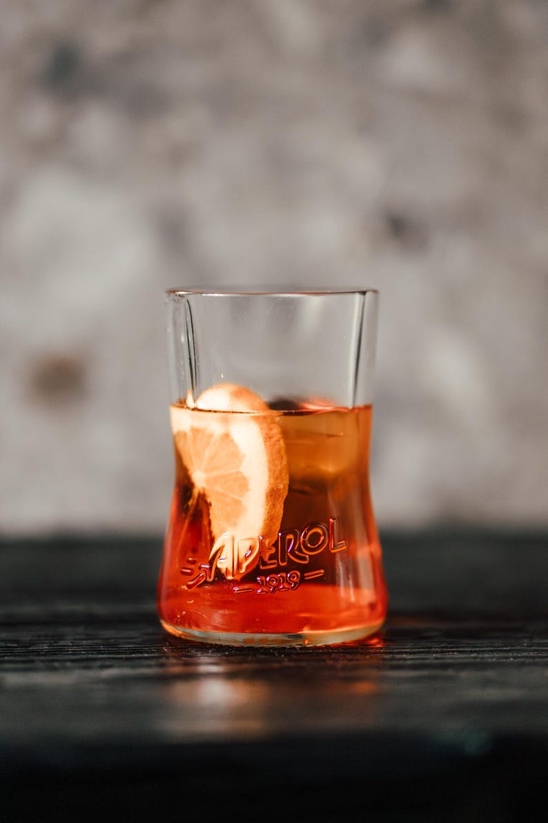 May include: A glass of Aperol Spritz with a slice of orange. The glass is clear and has the word "Aperol" printed on it. The drink is a bright orange color.