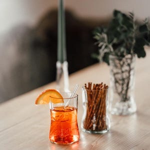 May include: A glass of orange juice with a slice of orange on the rim, a glass jar filled with pretzel sticks, and a lit candle in the background.