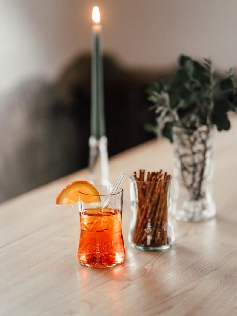 May include: A glass of orange juice with a slice of orange on the rim, a glass jar filled with pretzel sticks, and a lit candle in the background.