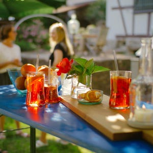 May include: A table setting with four glasses of orange juice, a bowl of oranges, and a vase of red flowers. The table is blue and has a wooden board on top. There is a candle on the table.