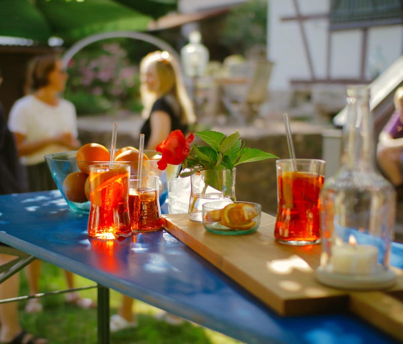 May include: A table setting with four glasses of orange juice, a bowl of oranges, and a vase of red flowers. The table is blue and has a wooden board on top. There is a candle on the table.