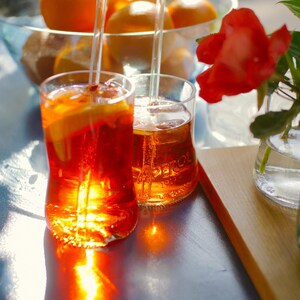 May include: Two glasses of Aperol spritz with orange slices and ice cubes. The glasses are on a blue table with a wooden surface in the background. There is a bowl of oranges in the background.