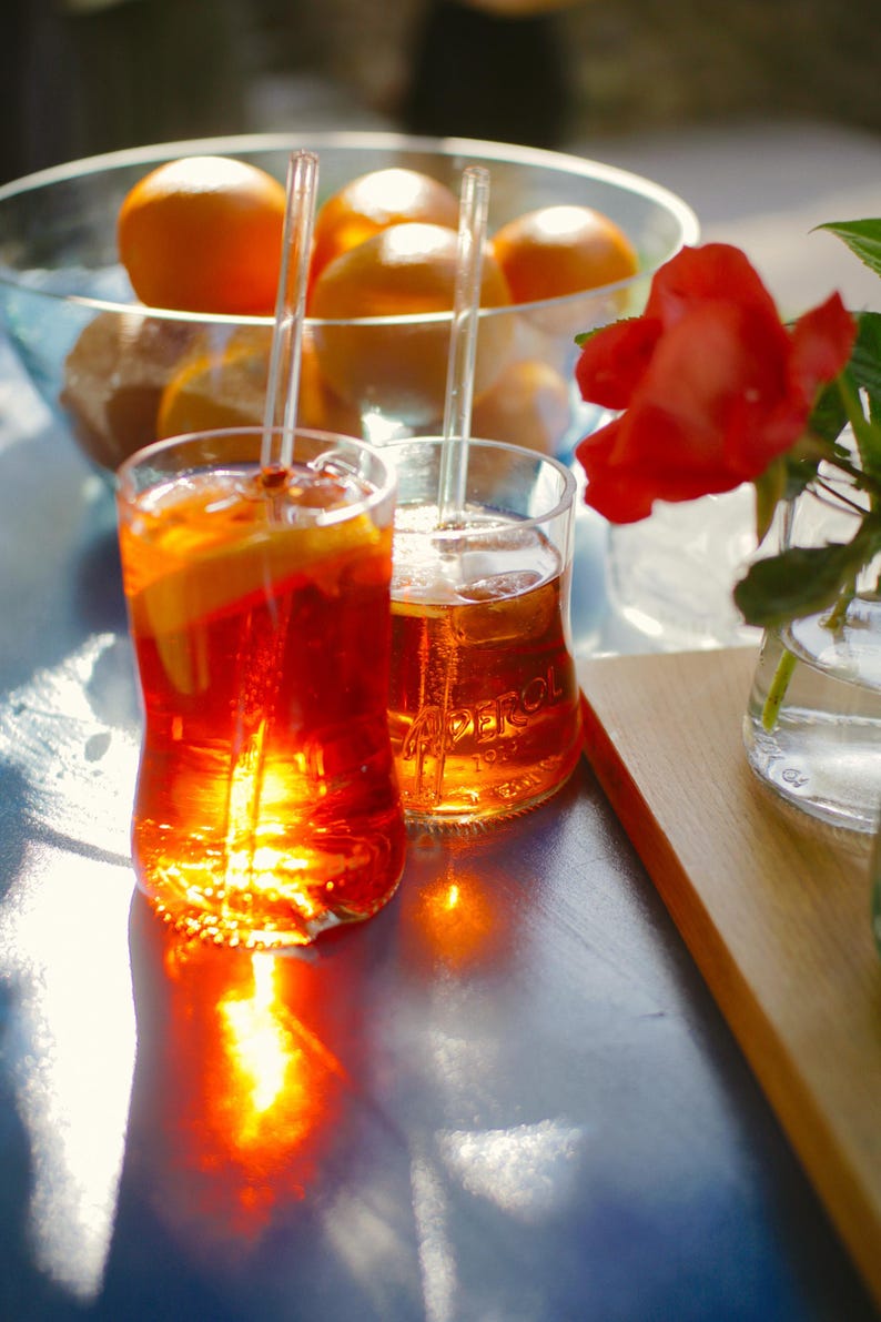 May include: Two glasses of Aperol spritz with orange slices and ice cubes. The glasses are on a blue table with a wooden surface in the background. There is a bowl of oranges in the background.