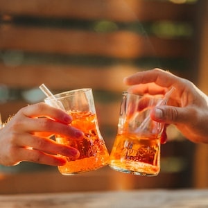 Könnte beinhalten: Zwei klare Gläser, gefüllt mit einem orangefarbenen Getränk, werden zum Anstoßen erhoben. Die Gläser haben eine leichte Sanduhrform und enthalten Eis und einen Strohhalm. Das Wort "Aperol" ist auf dem Glas zu sehen. Der Hintergrund ist eine verschwommene Holzoberfläche.