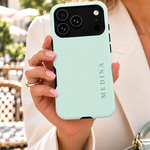 Könnte beinhalten: Eine mintgrüne Smartphone-Hülle mit schwarzem Kameramodul und dem vertikal aufgedruckten Wort MEDINA in Schwarz. Die Hülle hat eine glatte, matte Oberfläche und präzise Aussparungen für die Kameralinsen und den Blitz auf der Rückseite.