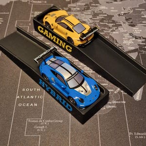 Könnte beinhalten: Zwei Spielzeug-Porsche-Rennwagen, einer gelb mit schwarzen Streifen und einer blau mit silbernen Streifen, werden in schwarzen Boxen präsentiert. Auf der Box des gelben Autos steht "GAMING", und auf der des blauen Autos "HYBRID".
