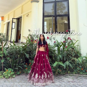 May include: A woman wearing a maroon lehenga, a traditional Indian outfit, stands in front of a building. The lehenga features intricate gold embroidery. The image includes the text "the lehenga store".