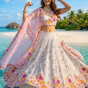 May include: A white and floral embroidered lehenga with a matching cropped top and a sheer pink dupatta. The outfit features colorful floral embroidery and is worn on a wooden dock near the ocean.