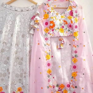 May include: Two-piece formal wear set. A white skirt with floral embroidery hangs next to a pink top and sheer shawl, also with floral embroidery. The top has a V-neck and tassel details. The floral embroidery includes pink, orange, and yellow.