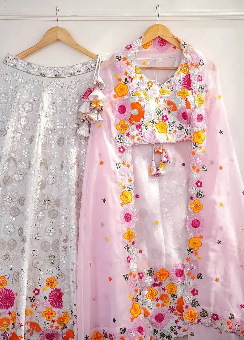 May include: Two-piece formal wear set. A white skirt with floral embroidery hangs next to a pink top and sheer shawl, also with floral embroidery. The top has a V-neck and tassel details. The floral embroidery includes pink, orange, and yellow.