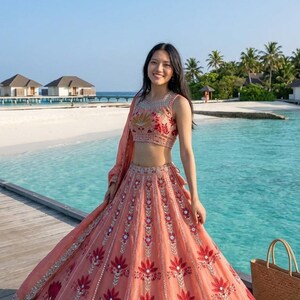 Puede incluir: Un lehenga bordado sin mangas de color melocotón con un dupatta a juego. La falda presenta grandes diseños florales rojos e intrincados adornos plateados. El atuendo se usa en un muelle de madera con un fondo de playa tropical.